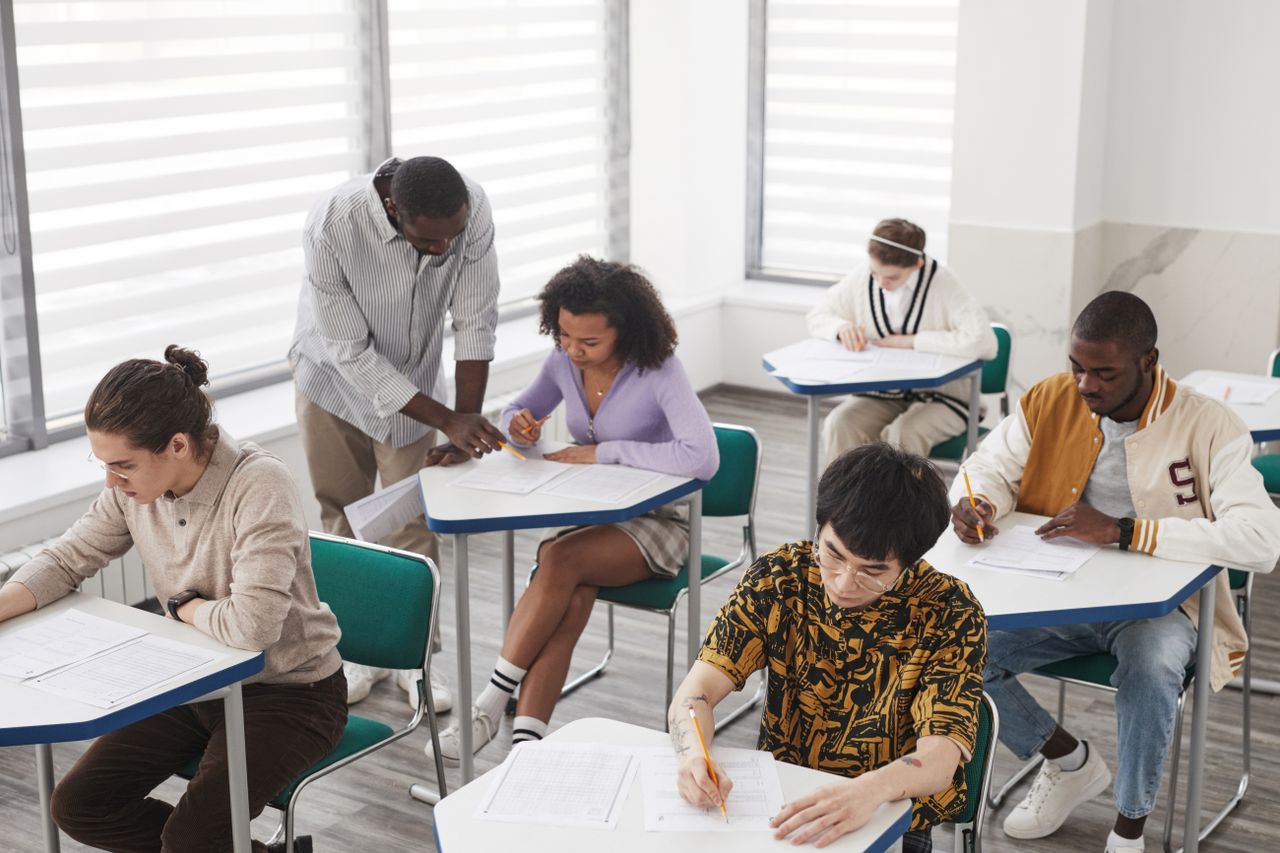 gallery-1 A diverse group of college students taking an exam, supervised by a teacher, in a classroom setting.